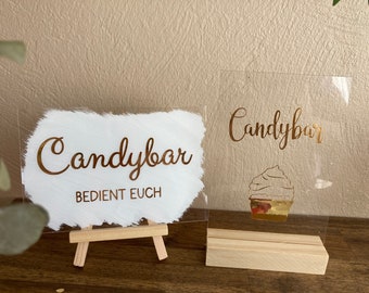 Puede incluir: Dos letreros de acrílico con letras doradas que dicen "Candybar". El letrero de la izquierda tiene un fondo blanco con el texto "BEDIENT EUCH" debajo de la palabra "Candybar". El letrero de la derecha tiene un fondo transparente con una ilustración de cupcake dorada. Ambos letreros están en soportes de madera.