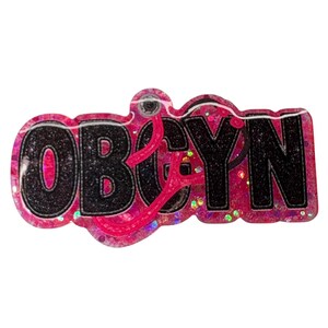 May include: A pink and black OBGYN badge with a stethoscope design. The letters "OBGYN" are in black with a glittery black outline, set against a pink background with glitter accents. A pink stethoscope is overlaid on the letters.
