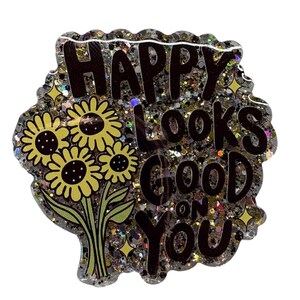 May include: A glittery, resin-based pin with the phrase "HAPPY looks GOOD on YOU" in black lettering. The pin features a bouquet of yellow sunflowers with green stems and leaves. The edges are scalloped.