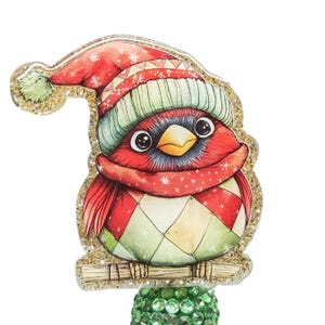 May include: A whimsical Christmas ornament featuring a red bird wearing a red and green hat and scarf. The bird has a green and red argyle pattern on its belly. The ornament is outlined with gold glitter and sits on a green, gem-studded base.