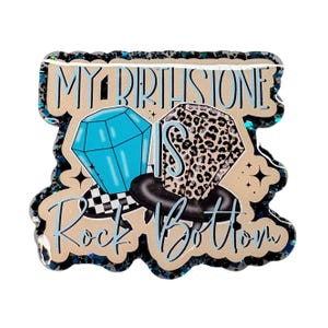 May include: A decorative pin with the text "MY BIRTHSTONE IS Rock Bottom". The pin features two diamond shapes, one blue and one with a leopard print pattern, and a black ring. The pin has a glittery border.