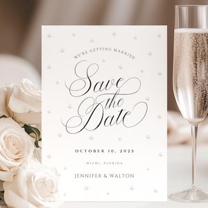 Puede incluir: Una tarjeta blanca de guardar la fecha con un diseño de perlas. El texto dice "We're getting married" y "Save the Date" en una fuente de escritura. La fecha es el 10 de octubre de 2025 y la ubicación es Miami, Florida. Los nombres Jennifer & Walton están en la parte inferior de la tarjeta.