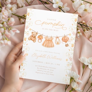 Pode incluir: Um cartão de convite retangular branco para chá de bebê com texto laranja que diz A LITTLE pumpkin IS ON THE WAY. O design inclui ilustrações de um ursinho de pelúcia, chocalho, vestido, babador e um balão de ar quente.