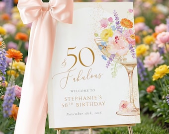 May include: A rectangular sign with a pale pink bow, featuring a watercolor illustration of a cocktail glass filled with colorful flowers. The text reads 50 & Fabulous, Welcome to Stephanie's 50th Birthday, and November 18th, 2026, in gold lettering.