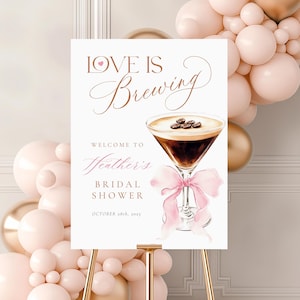 Può includere: Un cartello bianco su un cavalletto dorato con la scritta "Love is Brewing" e "Welcome to Heather's Bridal Shower October 28th, 2025". Illustrazione ad acquerello di un cocktail con chicchi di caffè e un fiocco rosa.