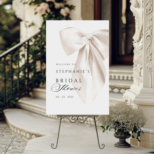 A vertical bridal shower welcome sign on a dark metal stand displayed outdoors on white steps next to a potted floral arrangement, with text reading Welcome to Stephanie's Bridal Shower