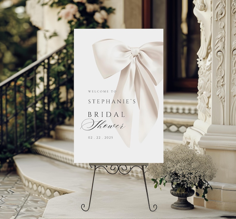 A vertical bridal shower welcome sign on a dark metal stand displayed outdoors on white steps next to a potted floral arrangement, with text reading Welcome to Stephanie's Bridal Shower