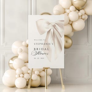 A vertical bridal shower welcome sign on a gold easel, featuring a large cream bow and text reading Welcome to Stephanie's Bridal Shower  surrounded by white and gold balloons.