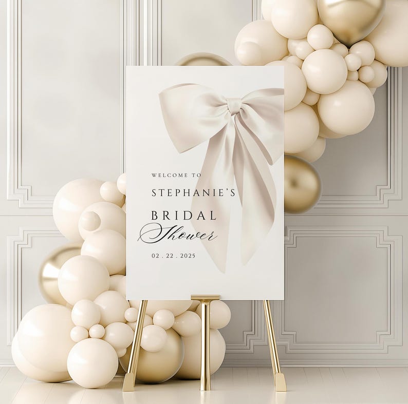 A vertical bridal shower welcome sign on a gold easel, featuring a large cream bow and text reading Welcome to Stephanie's Bridal Shower  surrounded by white and gold balloons.