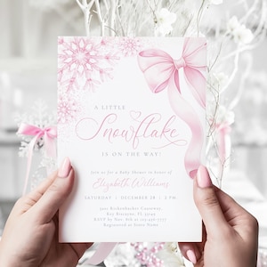 Peut inclure: Une invitation à une baby shower avec un motif de flocon de neige rose et un grand nœud rose. Le texte dit "A Little Snowflake Is On The Way !" et comprend les détails de l'événement. L'invitation est tenue par une personne.