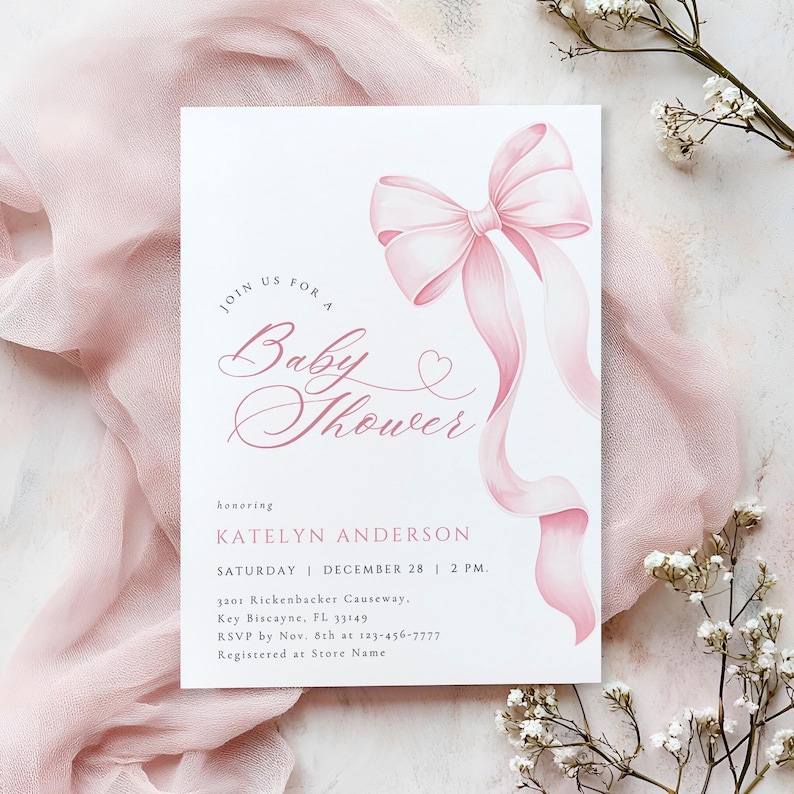 May include: A white baby shower invitation with a pink watercolor bow and flowing ribbons. The invitation includes the text "Join us for a Baby Shower" and event details in pink script. Floral accents complete the design.