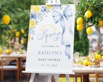 May include: A white easel displays a baby shower welcome sign. The sign features a blue bow and the text "Mama's Main Squeeze" and "Welcome to Katelyn's Baby Shower 02. 22. 2026". Lemon and floral accents decorate the sign.