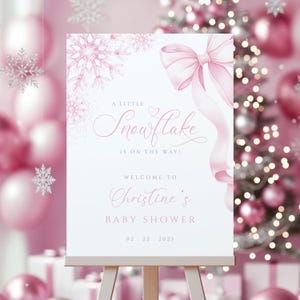 May include: A white baby shower sign with pink accents, featuring a snowflake design and a pink bow. The text reads "A Little Snowflake is on the Way!" and welcomes guests to Christine's baby shower on 02.22.2025.