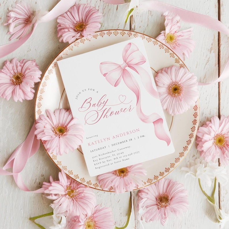May include: A baby shower invitation with a pink bow design, surrounded by pink gerbera daisies and ribbon. The invitation is on a decorative plate with a gold rim. The text reads "Join us for a Baby Shower".