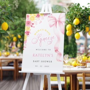 May include: A white welcome sign on an easel with pink text and a pink bow. The sign reads "Mama's Main Squeeze" and welcomes guests to Katelyn's baby shower on 02.22.2025. Floral and lemon accents decorate the top.