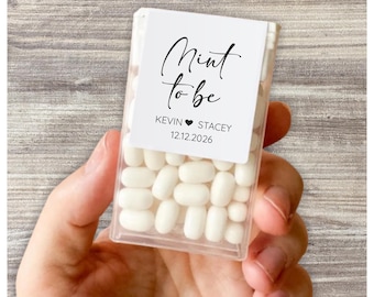 May include: A clear plastic box filled with white mints, held in a hand. The box has a white label with the words "Mint to be" in elegant script, along with the names Kevin and Stacey, and the date 12.12.2026.
