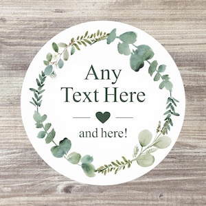 May include: A circular white sticker featuring a watercolour eucalyptus wreath design. The sticker has the text "Any Text Here" above a heart and the words "and here!" below. The background is a wood grain.