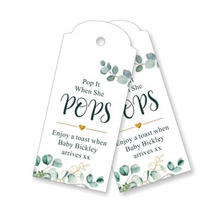 May include: Two white favor tags with gold and green eucalyptus designs. The text on the tags reads "Pop It When She Pops" and "Enjoy a toast when Baby Bickley arrives xx".