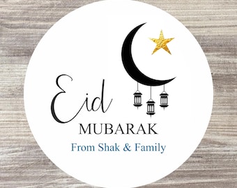 May include: White round sticker with a black crescent moon and a gold star. The text "Eid Mubarak" is written in black cursive script. Below the text is "From Shak & Family" in blue text.