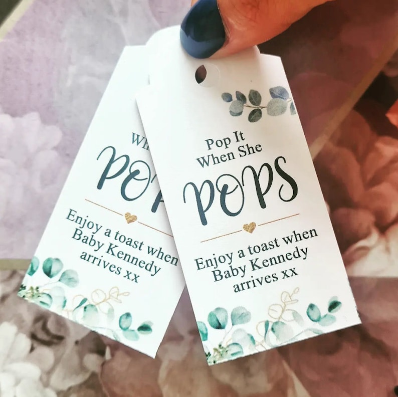 May include: Two white tags with a hole at the top. The tags have a green and blue floral design with gold text. The text on the tags reads "Pop It When She Pops" and "Enjoy a toast when Baby Kennedy arrives xx".