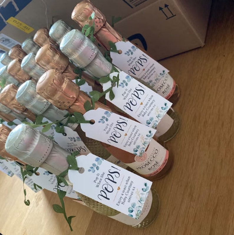 May include: A group of small champagne bottles with personalized tags that say "Pop It When She Pops!" and "Enjoy a toast when Baby Courtney arrives xx". The tags are decorated with a watercolor floral design in shades of blue and green.