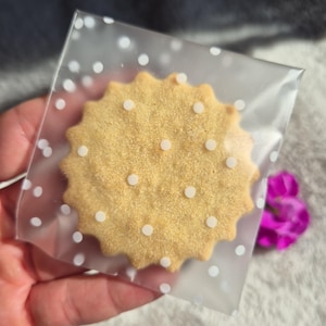 May include: A single, round, golden-brown biscuit with a scalloped edge and white dots, packaged in a clear plastic bag with white polka dots. The biscuit is held in a person's hand, with a blurred background of white fabric and purple flowers.
