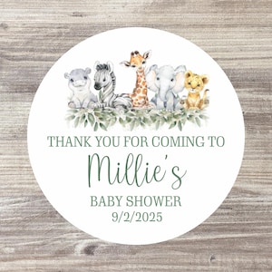 May include: A white circular sticker featuring a watercolour illustration of a hippo, zebra, giraffe, elephant, and lion. The text reads "Thank you for coming to Millie's Baby Shower 9/2/2025" in green script and block letters.