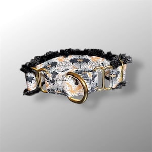 May include: A decorative dog collar featuring a repeating pattern of bats, sunbursts, and clouds in black and gold on a white background. The collar has gold-colored metal hardware and is trimmed with black fringe.