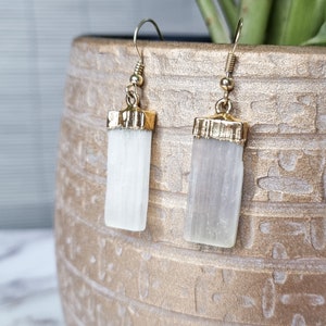 Può includere: Un paio di orecchini dorati con pietre di cristallo di selenite bianca. Gli orecchini sono di stile pendente con un gancio dorato.