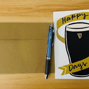 May include: A white card with the text "Happy Days" in black script. The card features a gold ribbon with a black glass of beer with a gold harp logo.