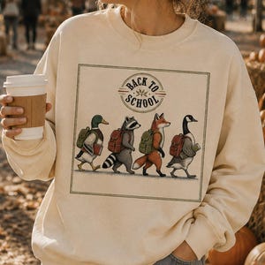Può includere: Una felpa girocollo color crema con una stampa grafica quadrata raffigurante un germano reale, un procione, una volpe e un'oca con zaini, che camminano in fila con la scritta "BACK TO SCHOOL" sopra, abbinata a un paio di jeans azzurri.