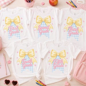 Könnte beinhalten: Fünf weiße T-Shirts mit kurzen Ärmeln und einer Grafik einer großen gelben Schleife über einem hellblauen Vichy-Karo-Wimpel. Jedes Shirt zeigt rosa Text mit First Grade, Second Grade, Third Grade, Fourth Grade oder Fifth Grade und kleinen Herzen.