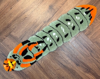 May include: A tufted rug shaped like a caterpillar with a segmented body in shades of sage green, black, and bright orange. The design features a rounded head with orange and black markings, a long body with black spots, and an orange tail tip.