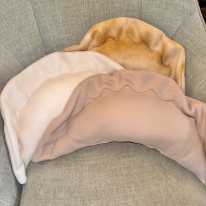 May include: Three plush dumpling-shaped pillows in varying shades of beige and tan. The pillows have a scalloped edge design, resembling the shape of a dumpling. They are arranged on a light green upholstered chair.
