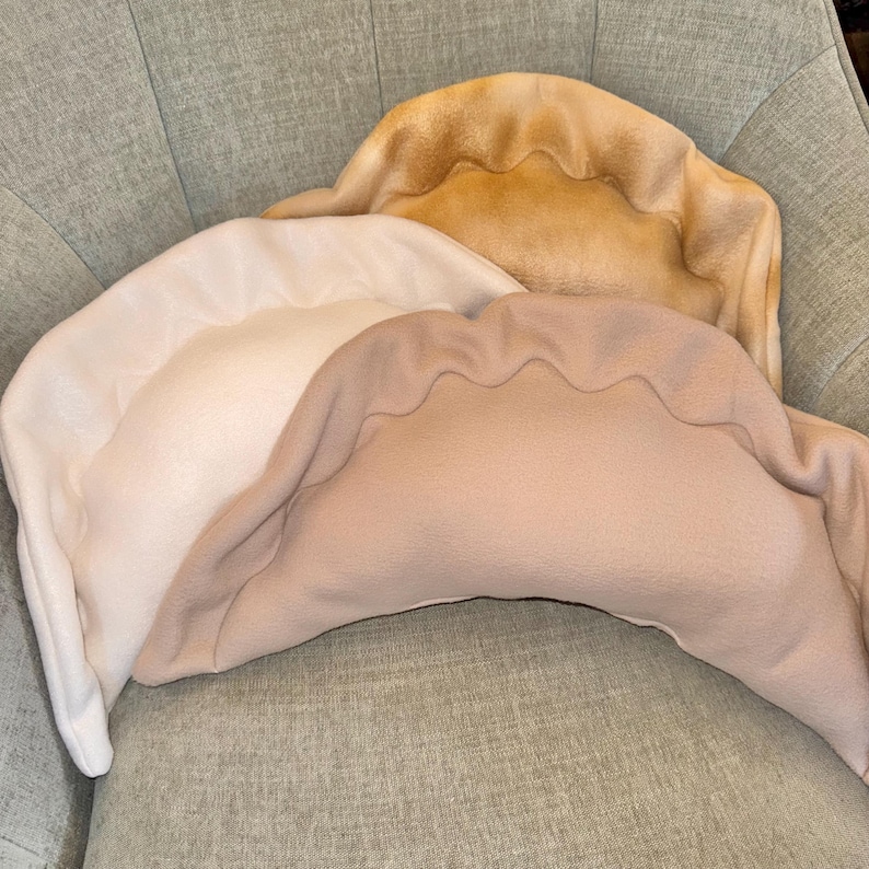 May include: Three plush dumpling-shaped pillows in varying shades of beige and tan. The pillows have a scalloped edge design, resembling the shape of a dumpling. They are arranged on a light green upholstered chair.