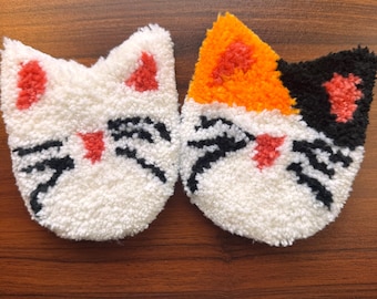 May include: Two white fuzzy cat-shaped coasters with embroidered faces. One coaster has black and orange fur, the other has black and red fur.