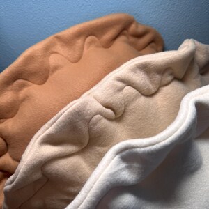 May include: Close-up of three fleece items in shades of tan, cream, and white. Each item has a scalloped edge, creating a decorative border. The soft texture and neutral colors suggest a cozy, handmade craft or home decor item.