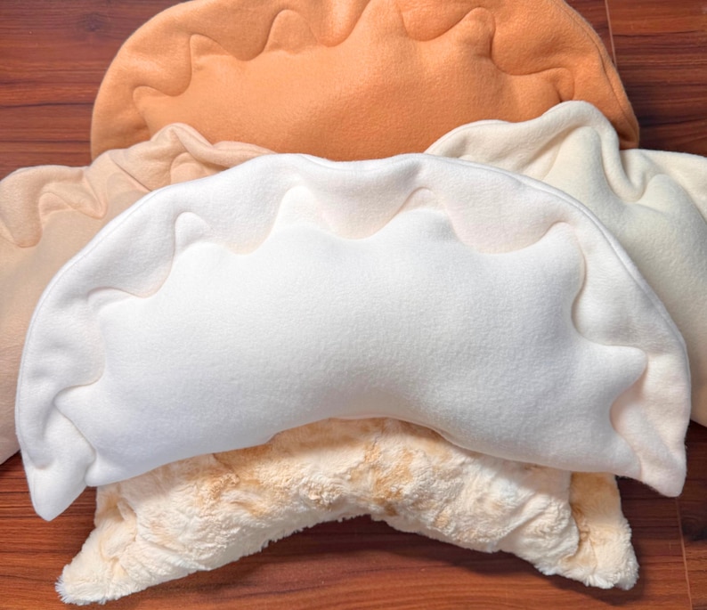 May include: A stack of plush dumpling-shaped pillows in various colors. The pillows are in shades of white, cream, and tan, with a textured, wavy edge design. The pillows are arranged on a wooden surface.