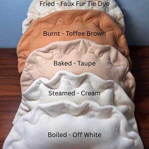 May include: A stack of plush, dumpling-shaped pillows in various colors. The pillows are labeled "Fried - Faux Fur Tie Dye", "Burnt - Toffee Brown", "Baked - Taupe", "Steamed - Cream", and "Boiled - Off White".