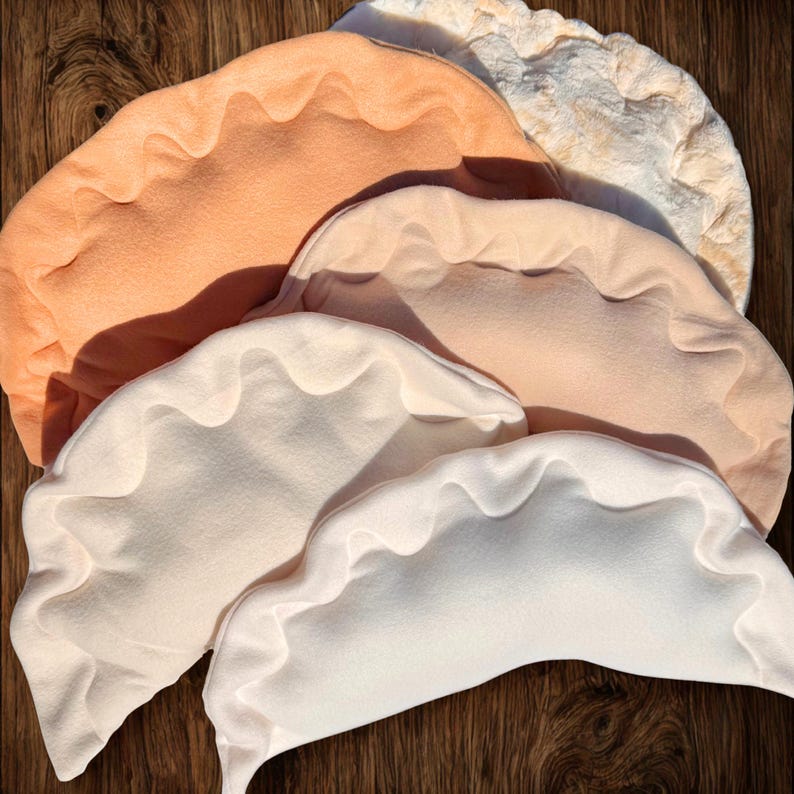 May include: A collection of plush, dumpling-shaped pillows in various colors, including peach, cream, and off-white. The pillows have a ruffled edge design and are arranged on a wooden surface.