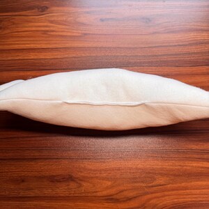 May include: A side view of a cream-colored pillow with a seam running along the center. The pillow is made of a soft, plush material and is resting on a dark brown wooden surface.
