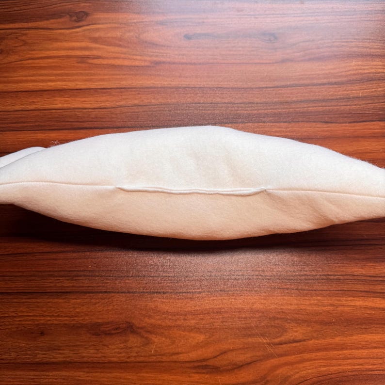 May include: A side view of a cream-colored pillow with a seam running along the center. The pillow is made of a soft, plush material and is resting on a dark brown wooden surface.