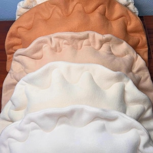 May include: A stack of plush, dumpling-shaped pillows in various neutral colors, including cream, tan, and brown. The pillows have a wavy, scalloped edge, and the top pillow is made of faux fur.