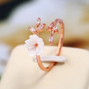 May include: A rose gold adjustable ring with a white flower and leaves accented with clear rhinestones.