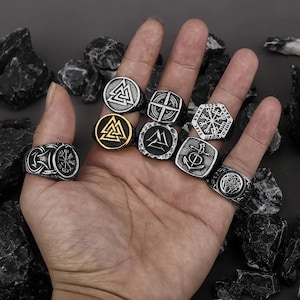 May include: A collection of eight silver and gold rings featuring various Viking symbols, including the Valknut, Vegvisir, and the anchor. The rings are worn on a hand.