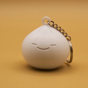 Pode incluir: Chaveiro branco impresso em 3D em forma de bolinho com um rosto sorridente.