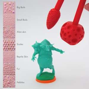 Könnte beinhalten: Eine grüne Plastik-Miniaturfigur eines Wesens mit drei Köpfen und einer strukturierten Oberfläche. Die Figur steht auf einem grünen Sockel mit orangefarbenem Rand. Zwei rote Plastikwerkzeuge mit strukturierten Oberflächen sind über der Figur zu sehen.