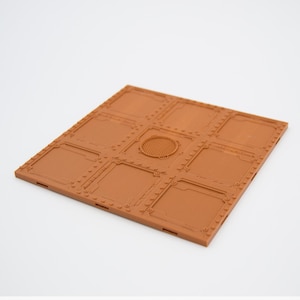 Puede incluir: Una baldosa cuadrada marrón texturizada con un patrón de cuadrícula y una ventilación circular en el centro. La baldosa está hecha de un material plástico duro y tiene un borde ligeramente elevado.