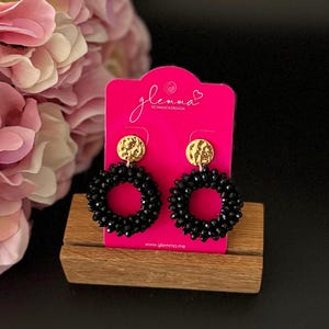 May include: Black beaded hoop earrings with gold textured disc accents. The earrings are displayed on a pink card with the text "glemma SCHMUCKDESIGN" and the website address "www.glemma.me".