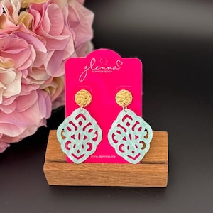 May include: A pair of light blue earrings with a gold-toned metal top and a delicate, intricate cutout design. The earrings are displayed on a pink card with the text "glemma Schmuckdesign" and "www.glemma.me".